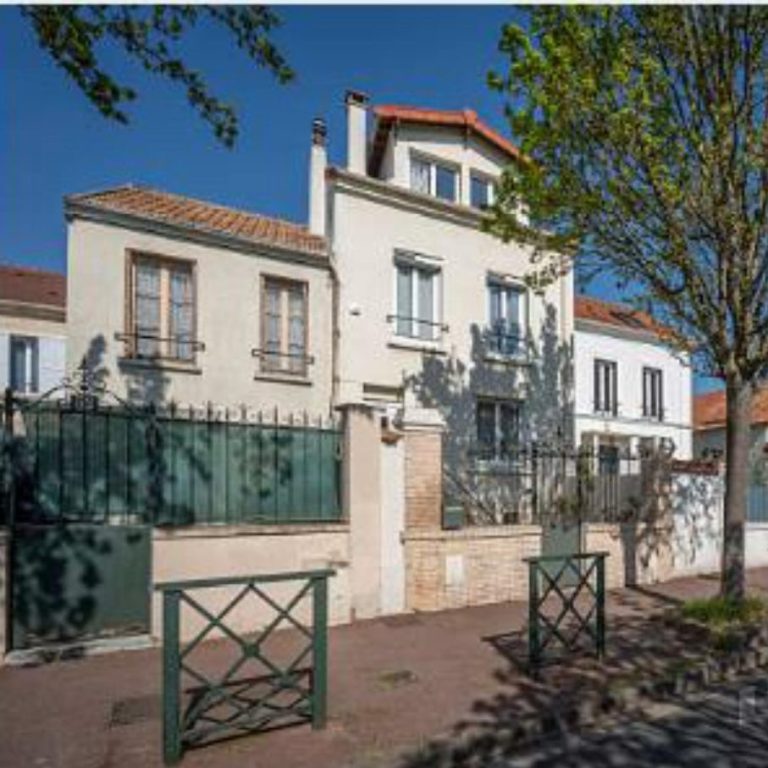 KITAROLL-BATIMENT ACHAT MAISON Maison blanche à deux étages avec balcons, entourée d'arbres et d'une clôture en fer.